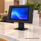A black tablet display stand with a mounted tablet on a marble counter in a modern office setting. The tablet screen displays a blue and purple abstract wallpaper.